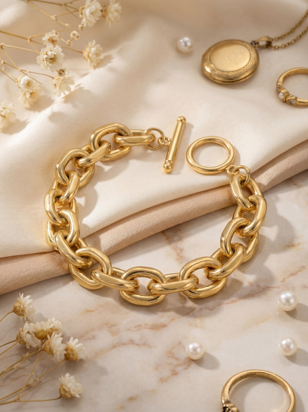 Gold chain bracelet on a marble surface with dried flowers and pearls.