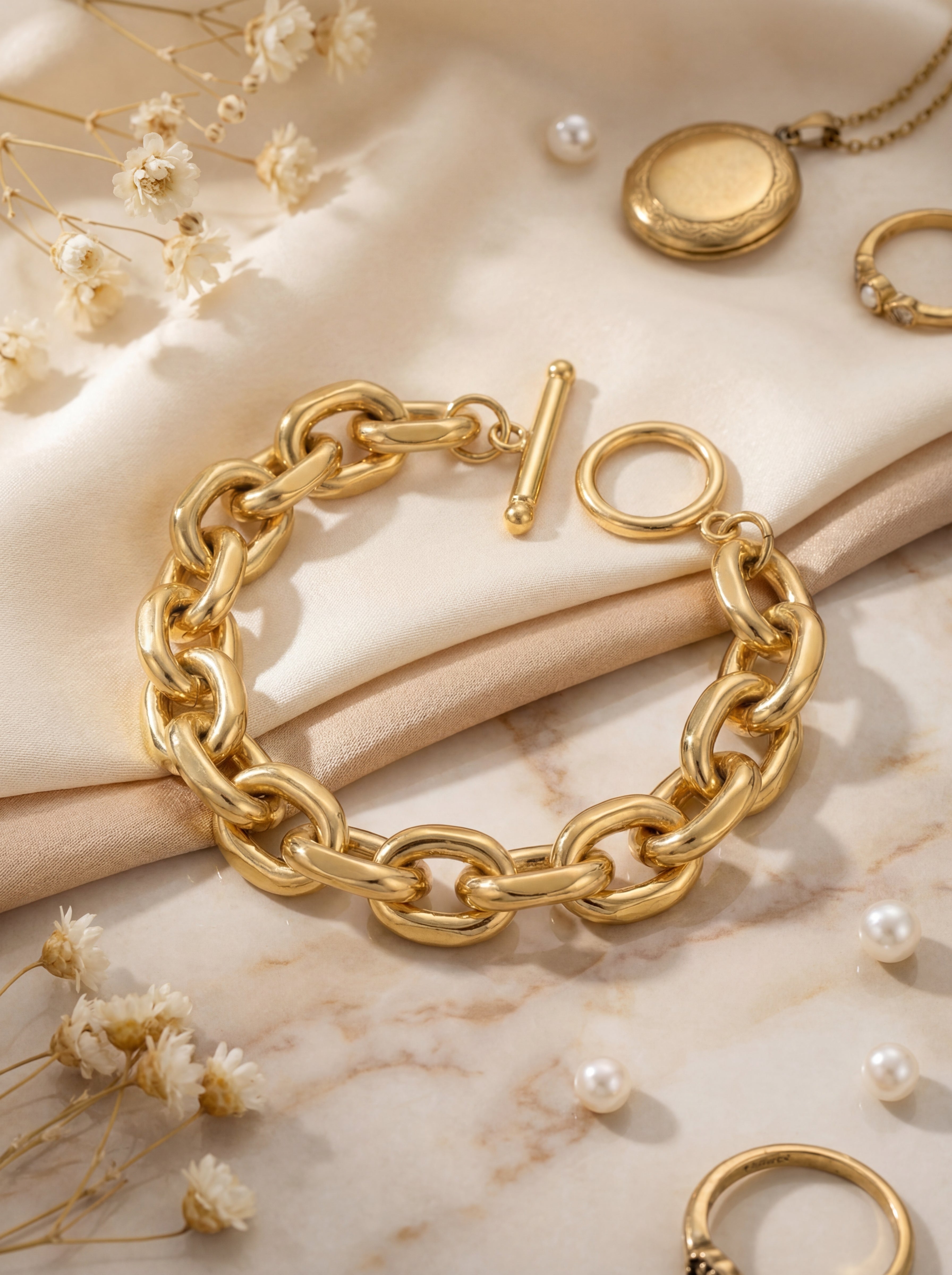 Gold chain bracelet on a marble surface with dried flowers and pearls.