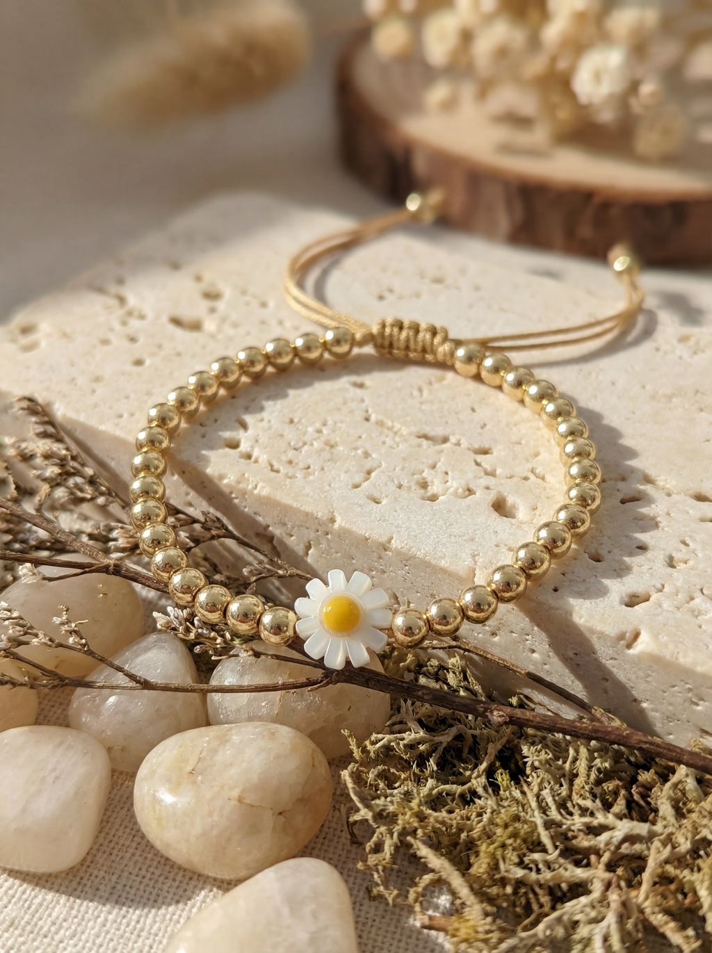 Gold beaded bracelet with a daisy charm on a natural stone surface with dried plants.