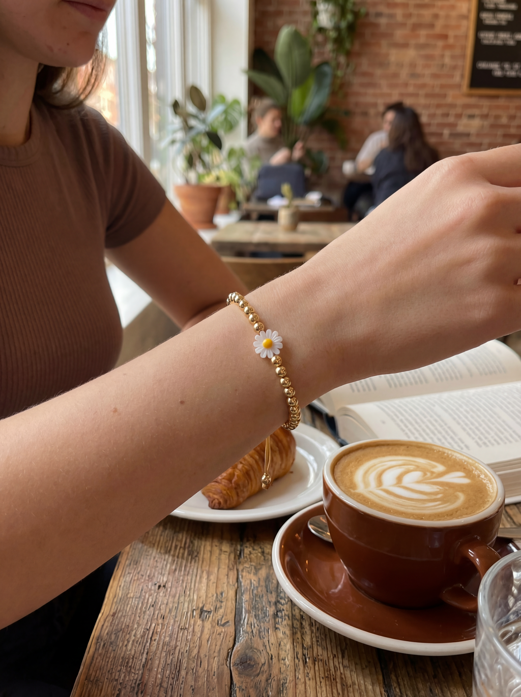 Person with a bracelet featuring a daisy charm at a cafe with a cup of coffee and pastry.