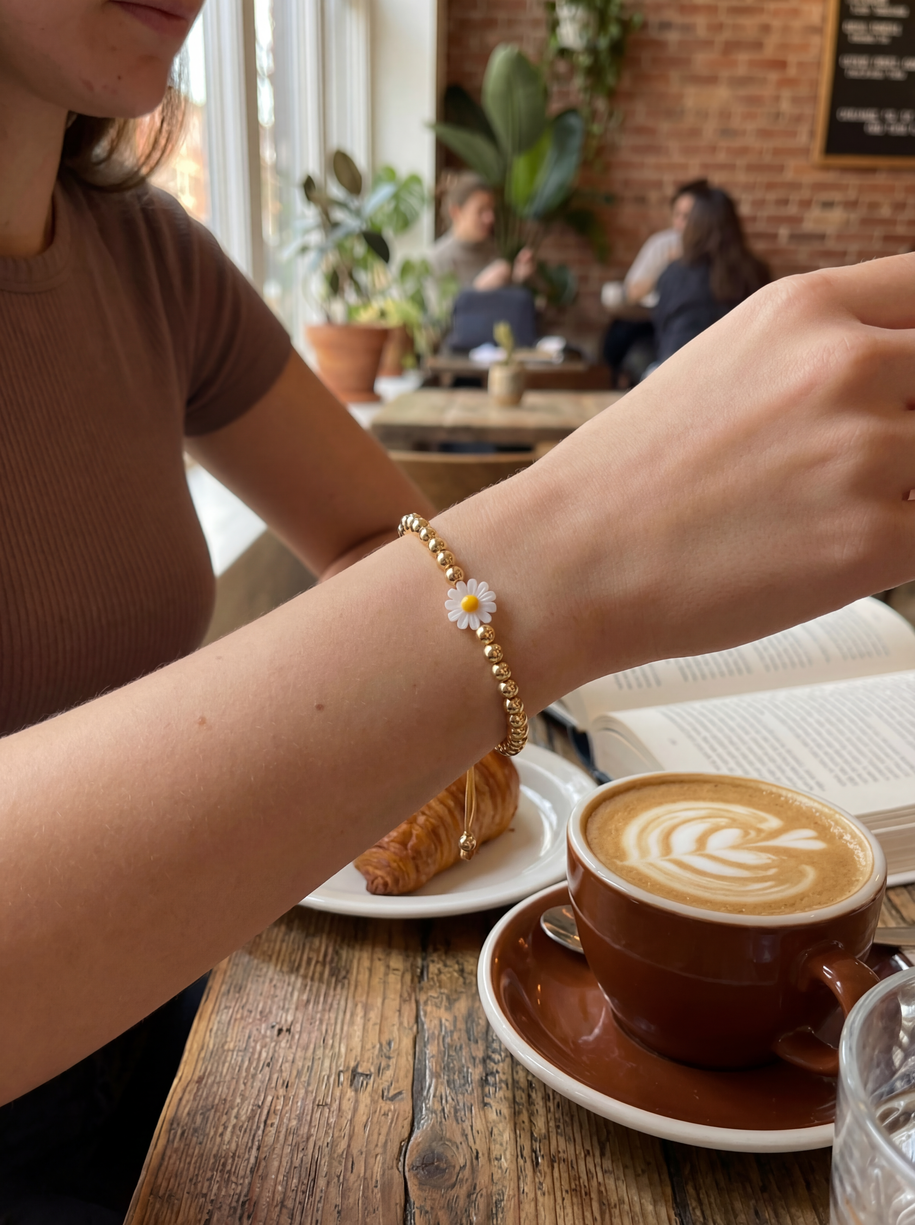 Person with a bracelet featuring a daisy charm at a cafe with a cup of coffee and pastry.
