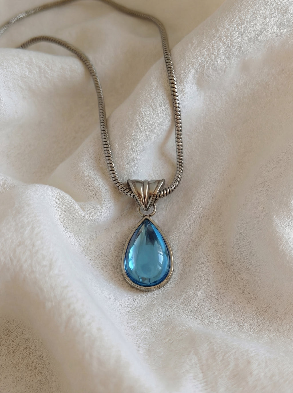Teardrop-shaped blue pendant on a silver chain against a textured white background