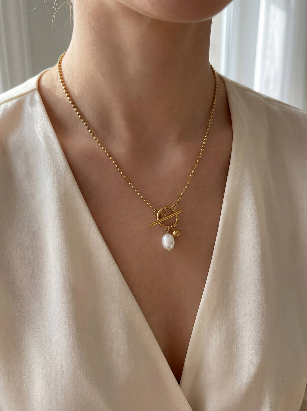 Gold necklace with a pearl pendant worn by a person in a white outfit.