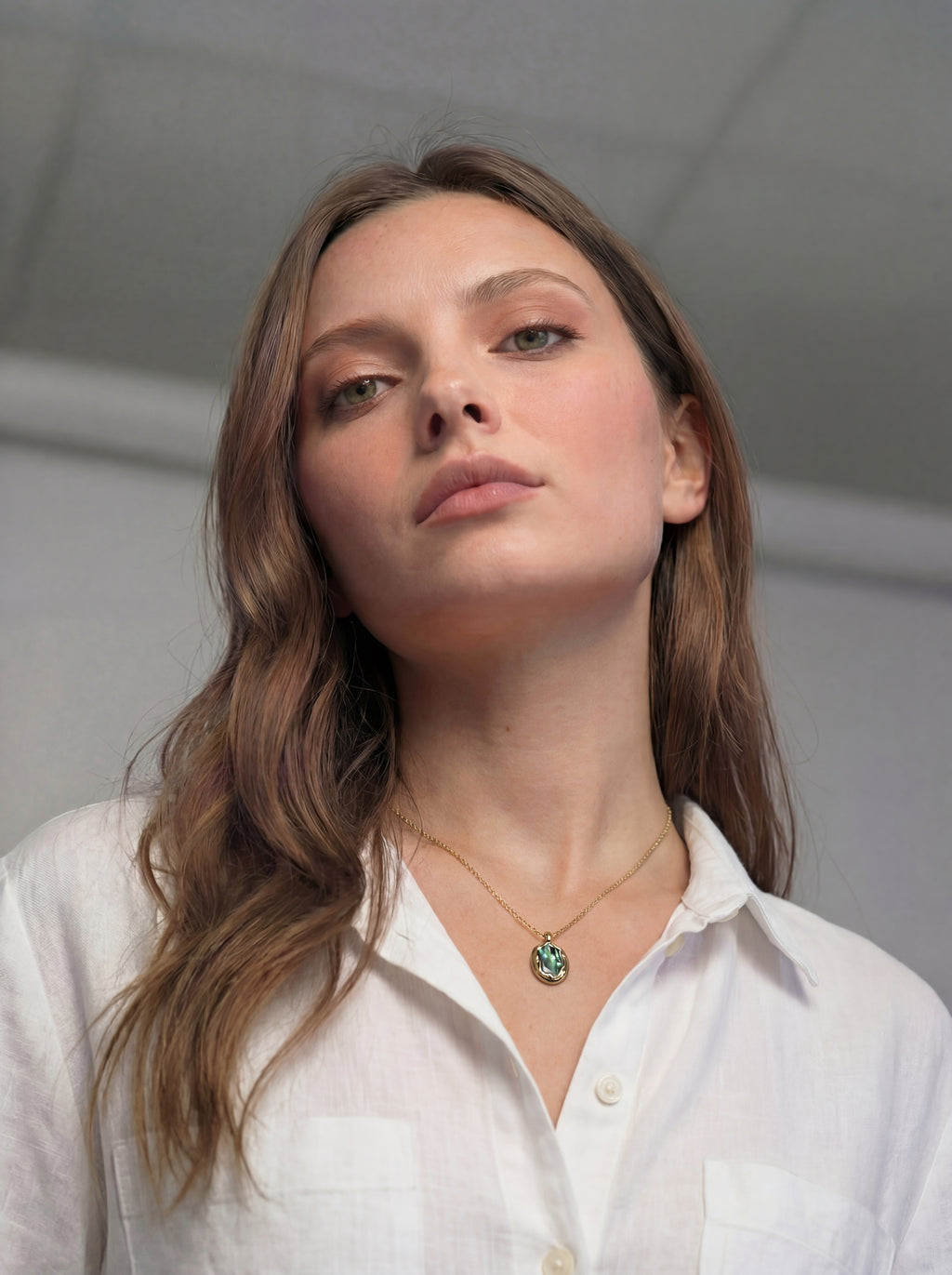 Woman wearing a white shirt and necklace with a green pendant against a neutral background