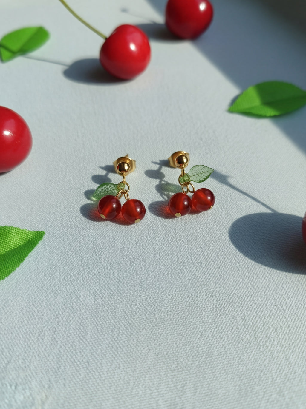 Cherry-themed earrings on a light surface with green leaves.