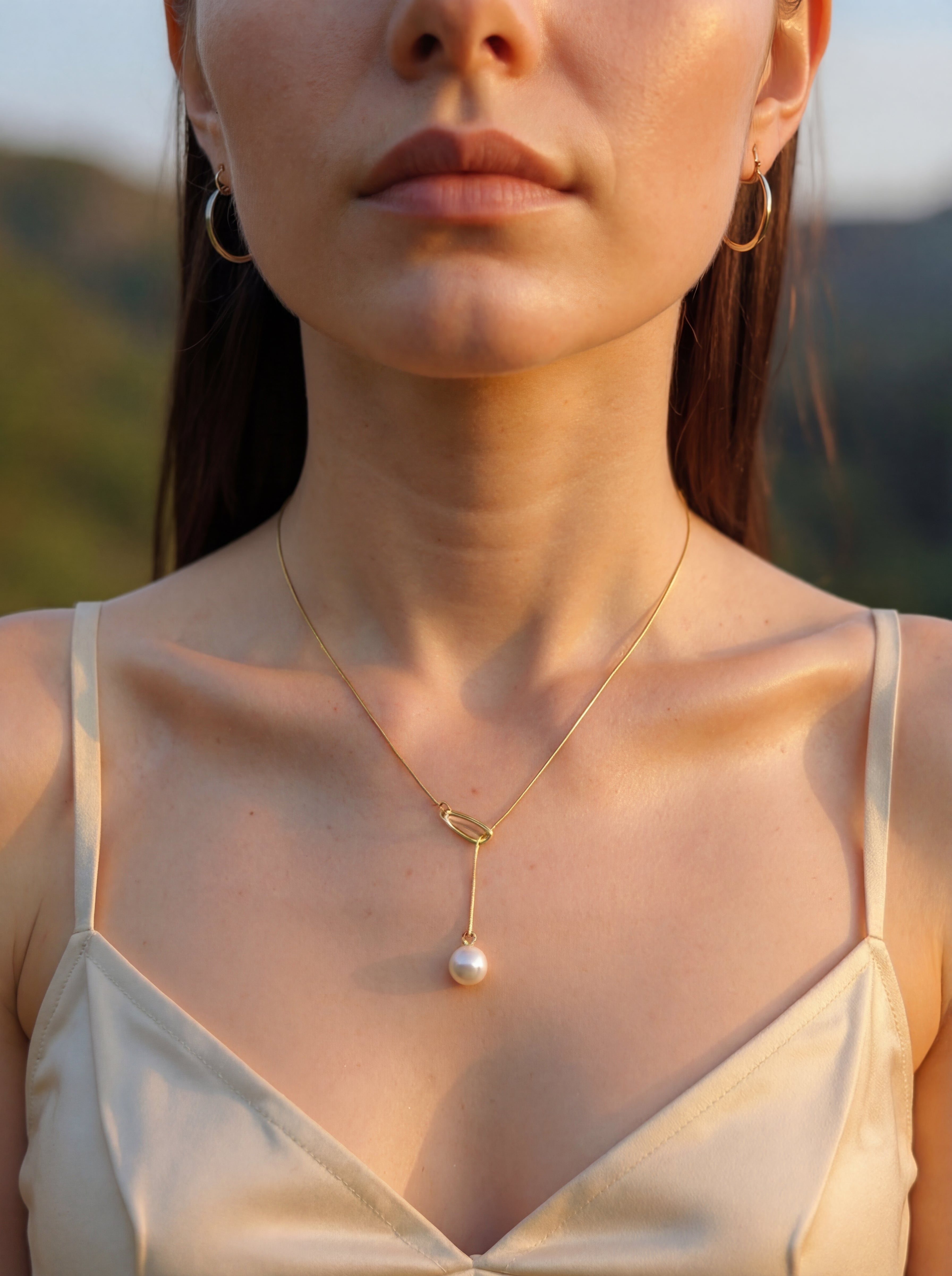 Close-up of a woman wearing a gold necklace with a pearl pendant against a blurred natural background.