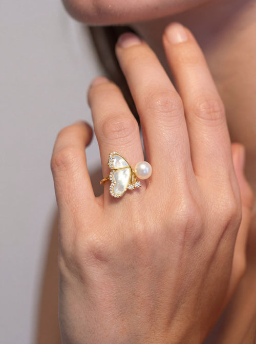Hand wearing a gold ring with a heart-shaped pearl on a neutral background