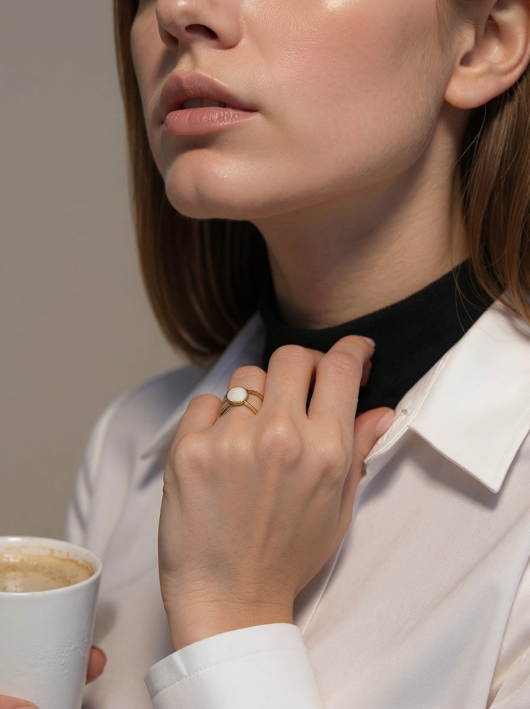 Woman adjusting her collar with a cup of coffee in hand, wearing a gold ring.