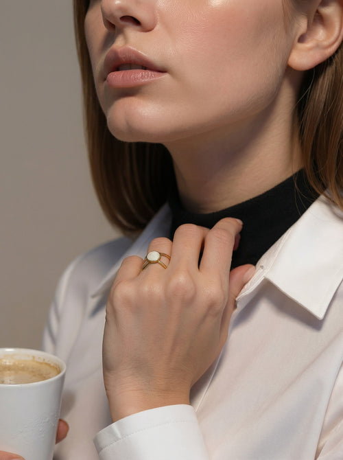 Woman adjusting her collar with a cup of coffee in hand, wearing a gold ring.