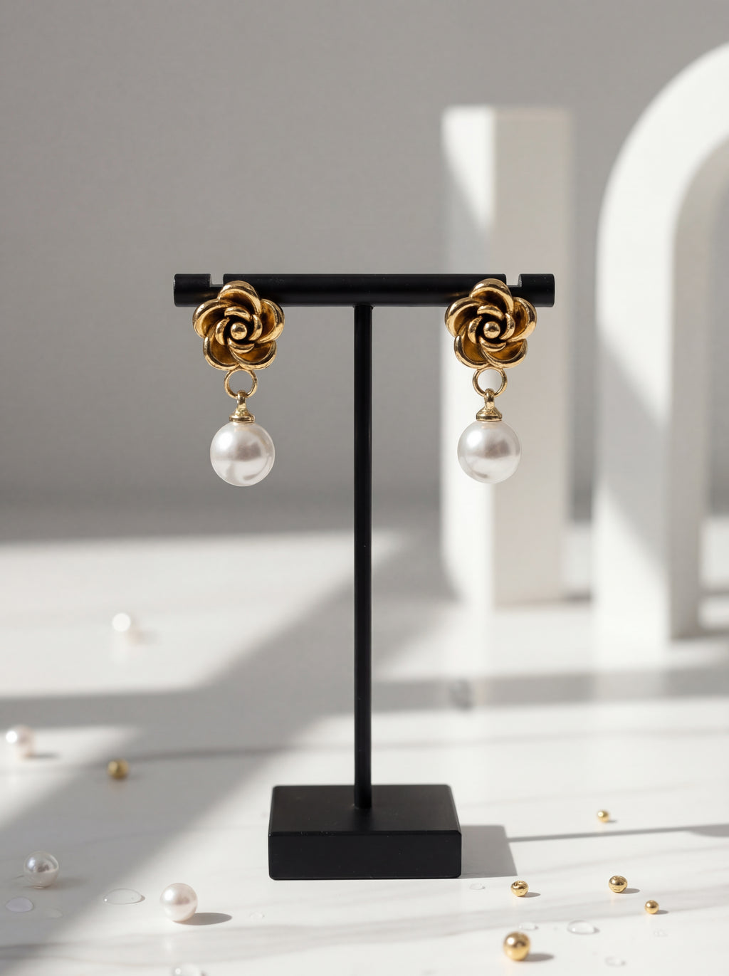 Gold and pearl earrings on a black stand with scattered pearls on a light surface.