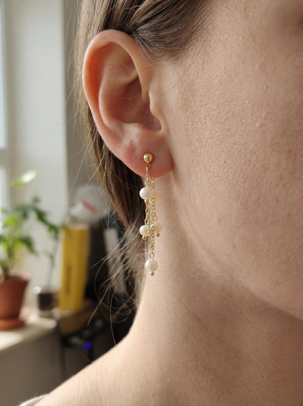 Close-up of a person wearing gold earrings with pearl drops, blurred indoor background.